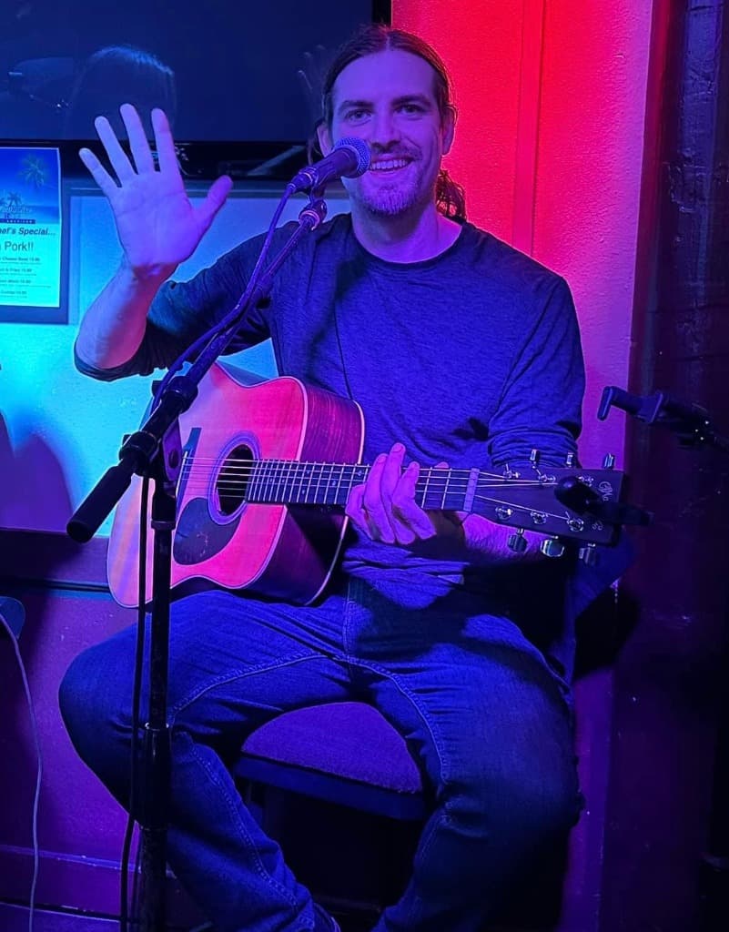 Luke Moore smiling and waving to the audience with acoustic guitar