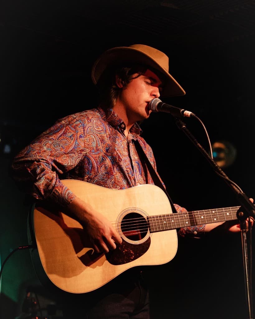 Luke Moore in western hat playing acoustic guitar into stage mic